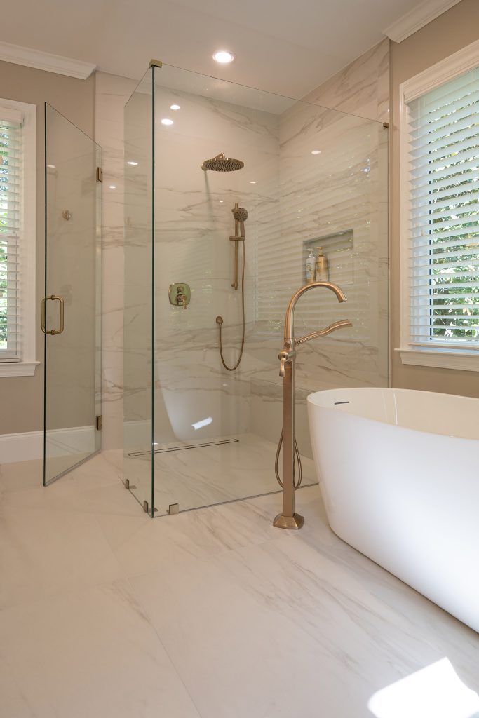White freestanding tub beside a marble-look glass shower with brushed-gold fixtures under a bright window.