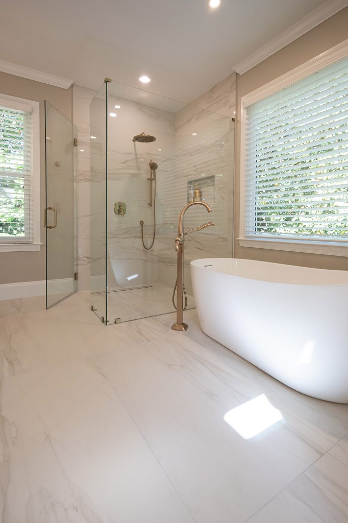 Freestanding soaking tub and marble-look glass shower with brushed-gold rain head and a built-in bench.