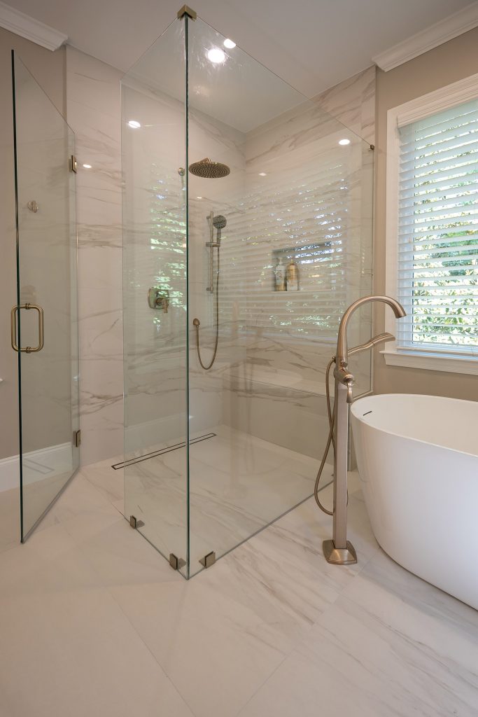 Corner glass shower in marble-look tile beside a freestanding tub with a brushed-gold floor-mount filler.