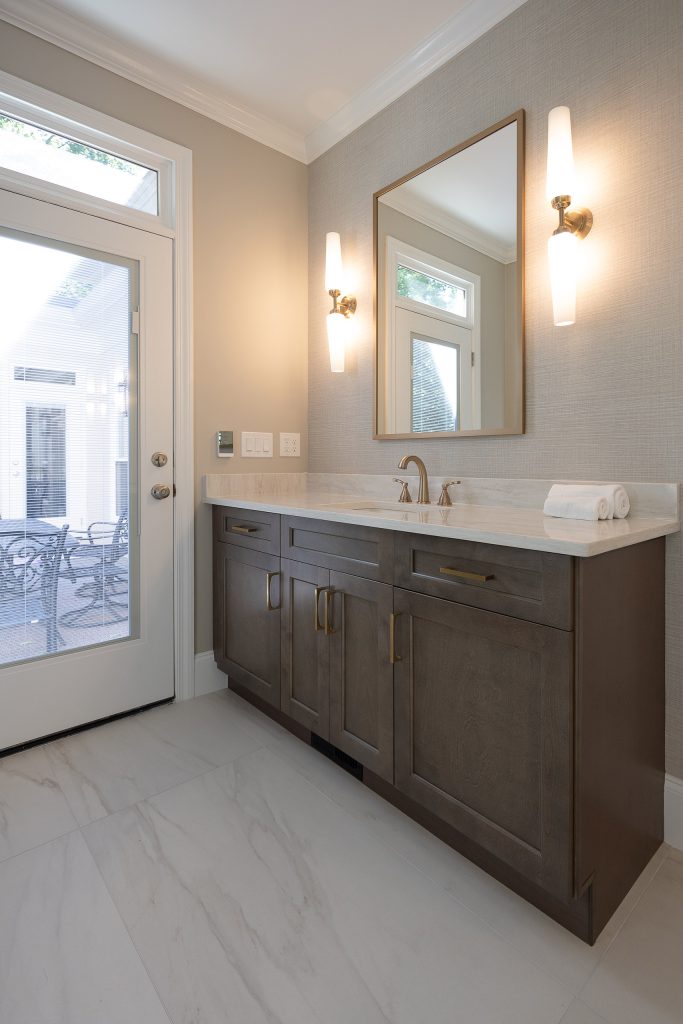 Single vanity with dark shaker cabinets and cream quartz top beside a glass patio door, under gold-framed mirror and sconces.