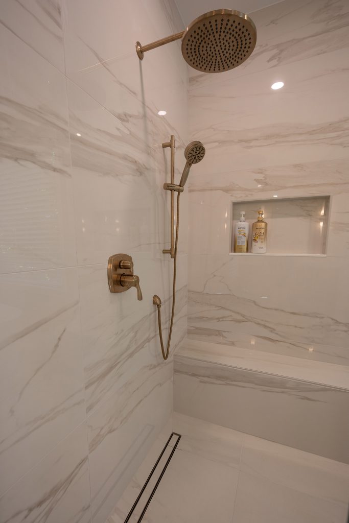 Marble-look shower interior with brushed-gold rain head, hand shower, a tiled bench, and a stocked niche.