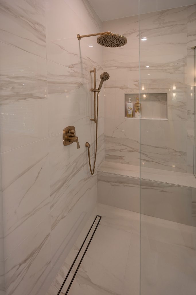Marble-look walk-in shower with brushed-gold rain head, hand shower, and a built-in niche behind frameless glass.