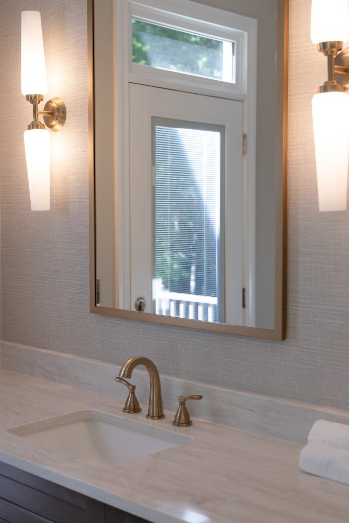 Vanity detail with a brushed-gold faucet, cream quartz counter, gold-framed mirror, and white-glass sconces on grasscloth walls.