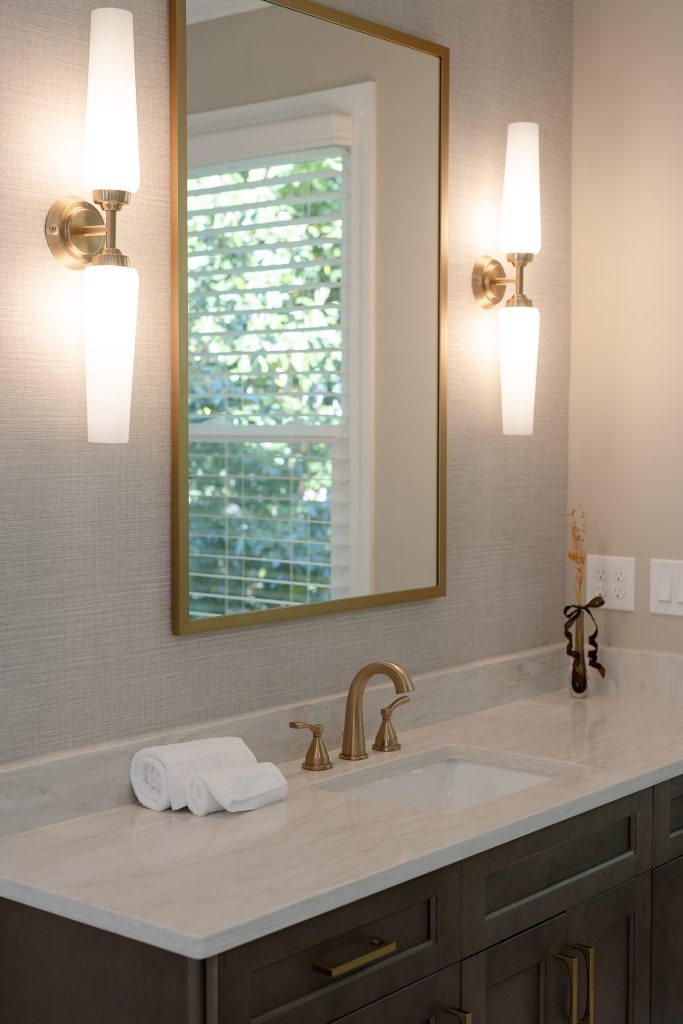 Vanity with dark-stained shaker cabinets, white quartz top, a gold-framed mirror, and cylindrical white-glass sconces.