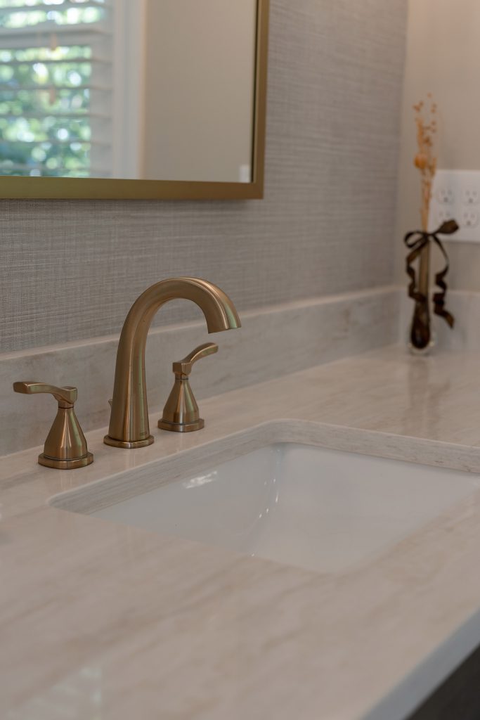 Champagne-bronze widespread faucet with lever handles on a soft cream quartz vanity top over a white undermount sink.