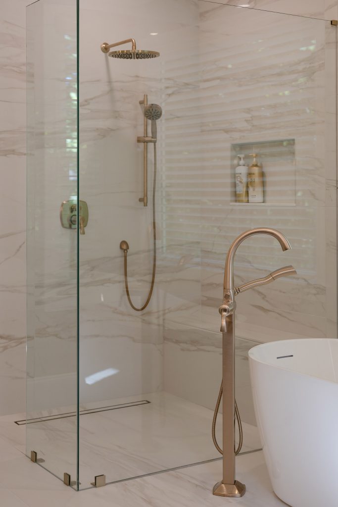 Marble-look walk-in shower with brushed-gold rain head and hand shower beside a white freestanding tub.