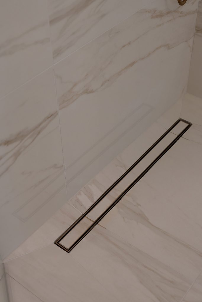 Linear shower drain set into a marble-look tile floor with veined walls above.