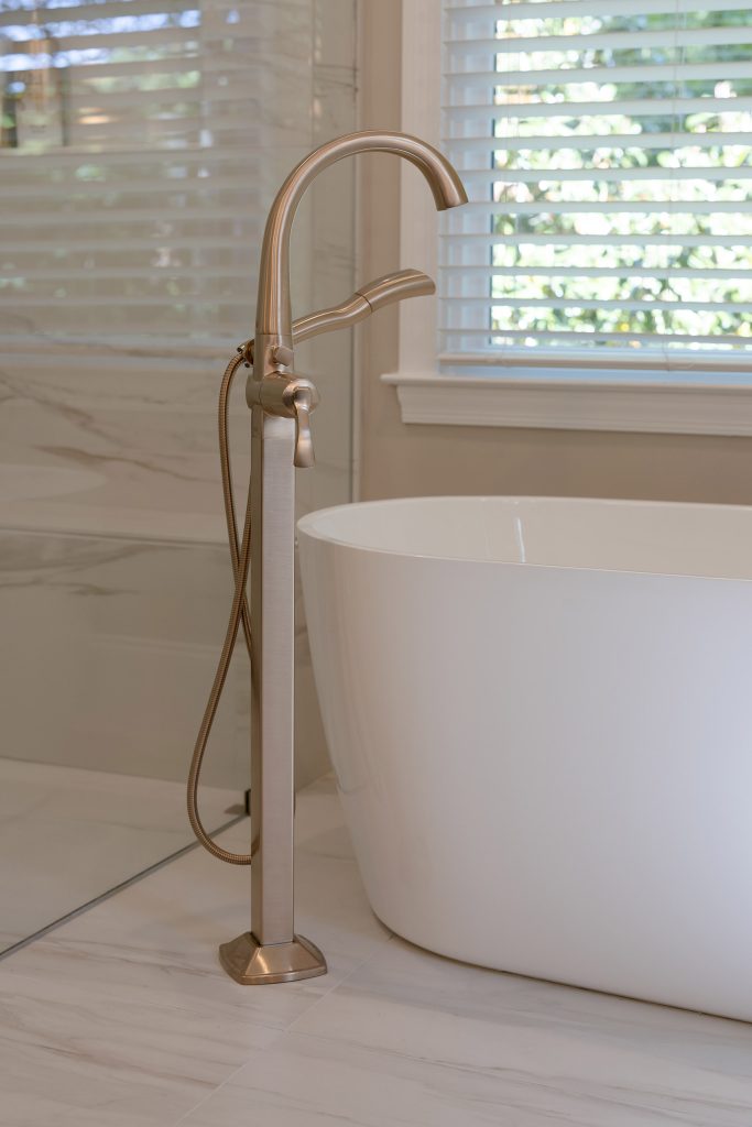 Brushed-gold floor-mounted tub filler and hand shower standing beside a white freestanding tub at a window.