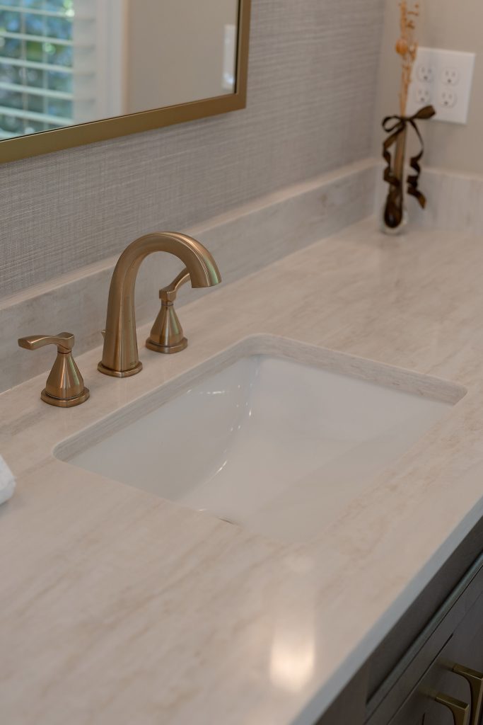 Vanity detail with a brushed-gold faucet on a cream quartz counter, white undermount sink, and grasscloth backsplash.
