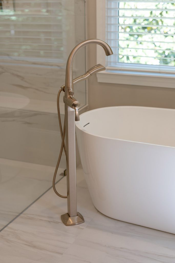 Curved white freestanding tub with a brushed-nickel floor-mount filler and hand shower beside a marble-look wall.