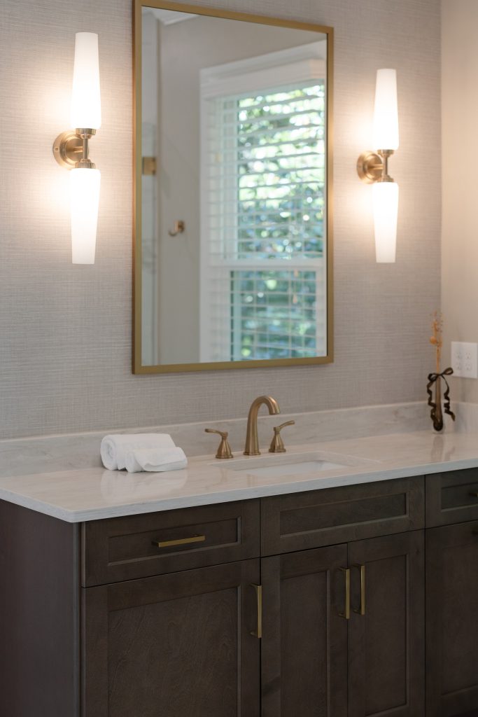 Dark shaker vanity with white quartz top, gold-framed mirror, and white-glass sconces on grasscloth-look walls.