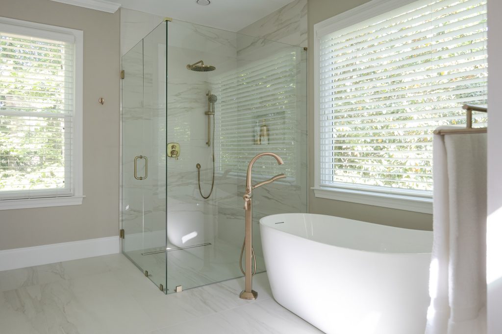 Marble-look corner shower with brushed-gold rain head and a white freestanding tub between two windows.