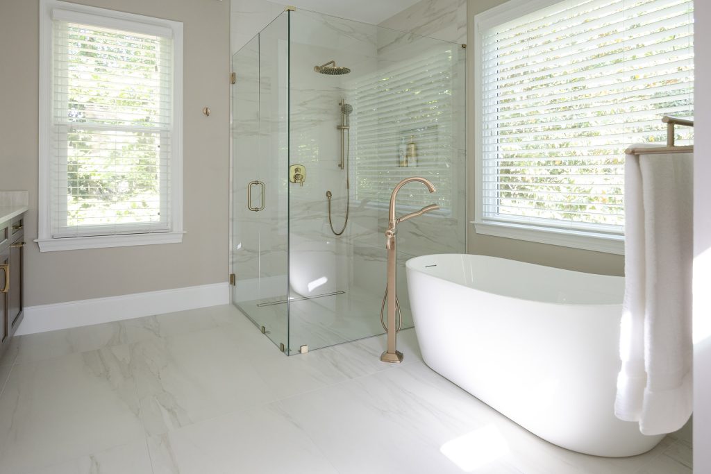 White freestanding tub beside a marble-look corner shower with brushed-gold filler and marble-look tile floors.