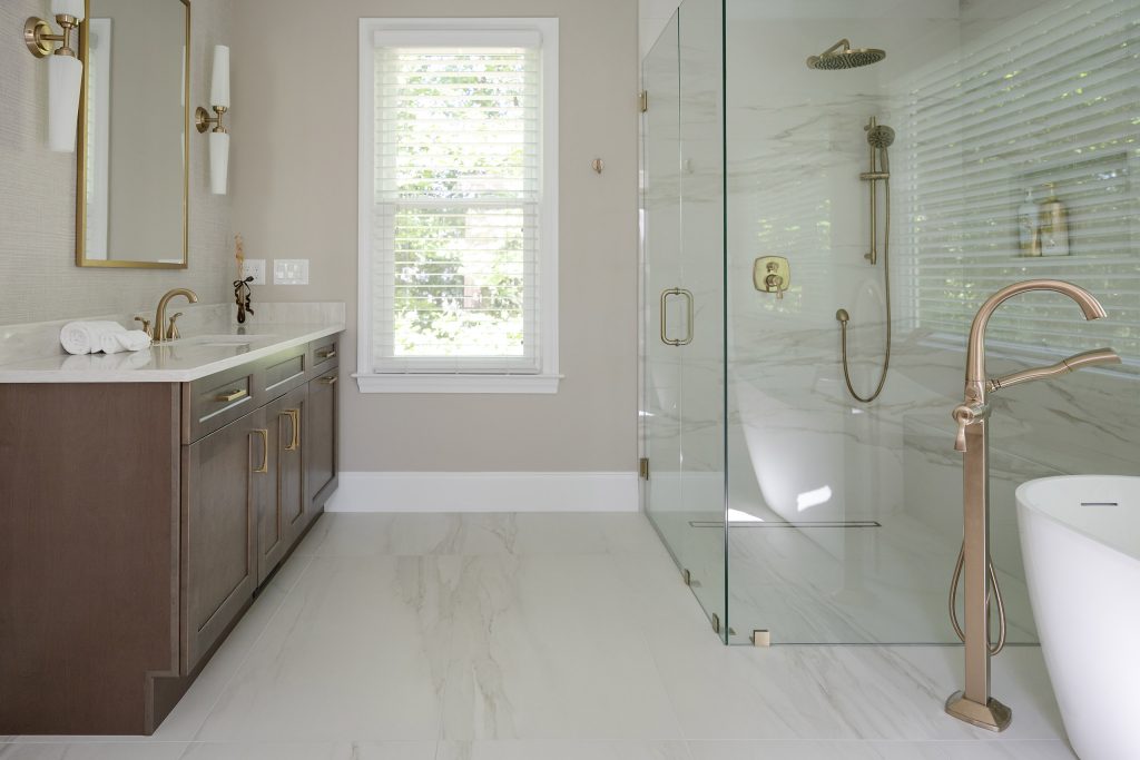 Master bath with a dark vanity, marble-look walk-in shower, and white freestanding tub with brushed-gold fixtures.