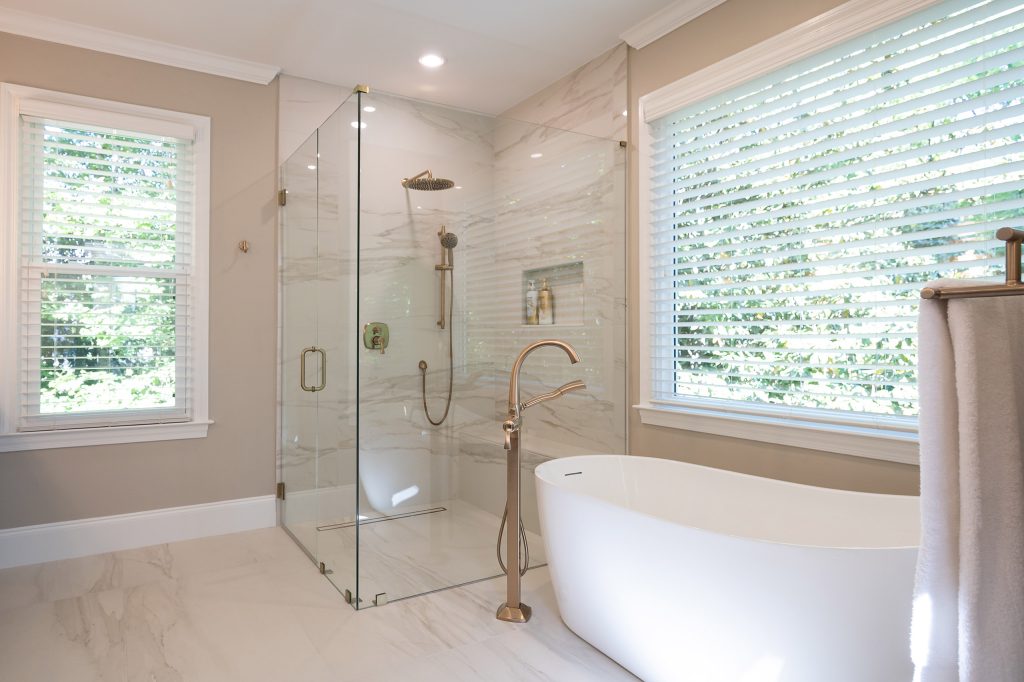 White freestanding tub and marble-look glass shower with brushed-gold rain head under two bright windows.