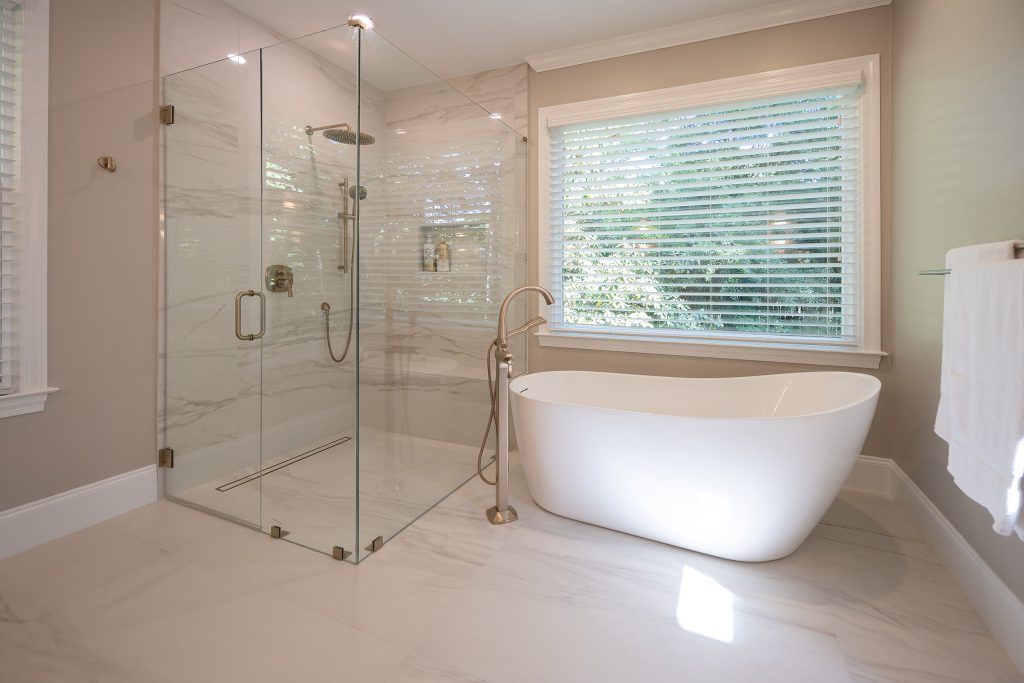 Corner glass shower in marble-look tile with brushed-gold fittings beside a white freestanding tub.