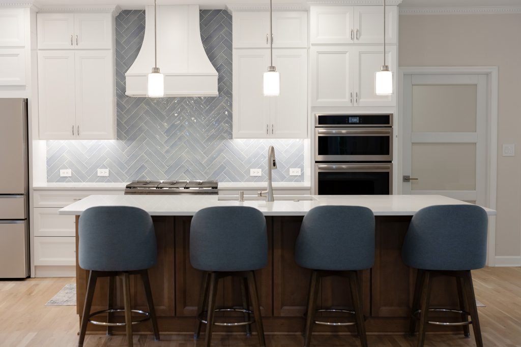 Open-concept kitchen with white inset cabinetry, a blue-grey herringbone tile backsplash, curved white hood, and a stained maple island seating four.