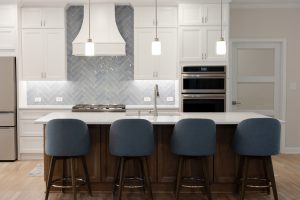 Open-concept kitchen with white inset cabinetry, a blue-grey herringbone tile backsplash, curved white hood, and a stained maple island seating four.