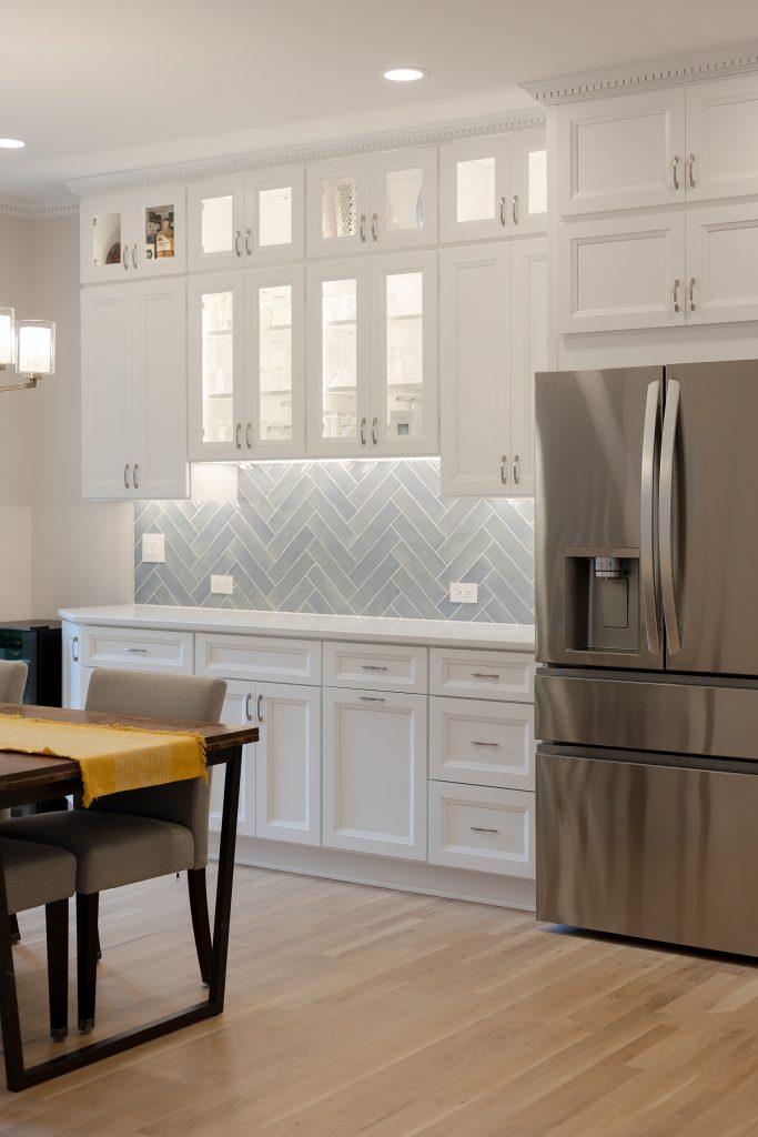Bar and beverage station where the cabinetry extends into the dining area, with illuminated glass-front uppers above the herringbone tile.
