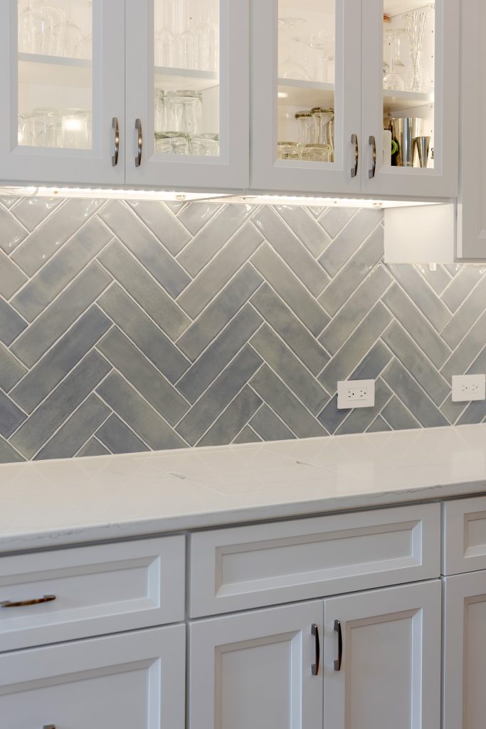 Illuminated glass-front cabinetry holding stemware above the herringbone backsplash and quartz counter of the beverage station.