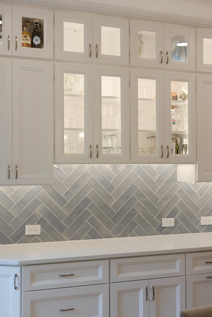 Glass-front upper cabinets lit from within, stocked with glassware above a blue-grey herringbone backsplash and white base cabinets.
