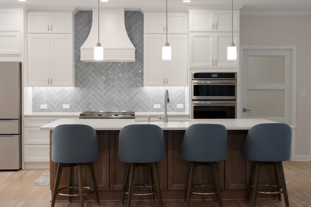 Remodeled kitchen seen across the island, with a curved white hood centred on a glossy herringbone backsplash and blue upholstered counter stools.