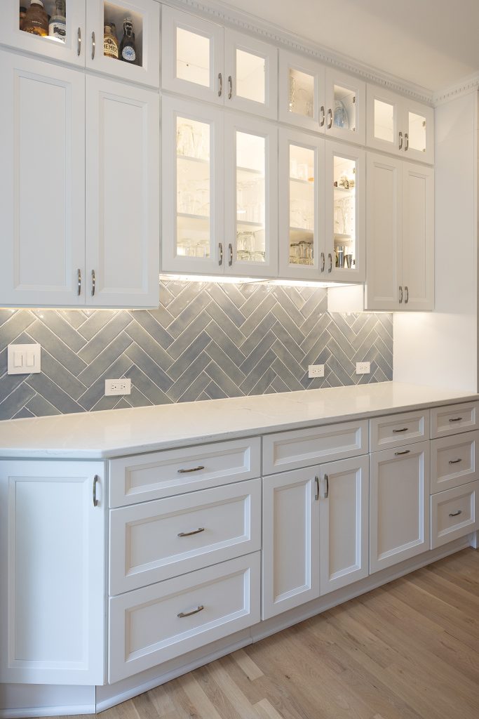 Full run of bar cabinetry with illuminated glass uppers, herringbone backsplash and white drawer bases on white oak flooring.