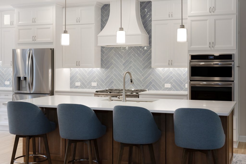 Angled view of the kitchen showing the stainless refrigerator, quartz-topped island and stacked double wall ovens.