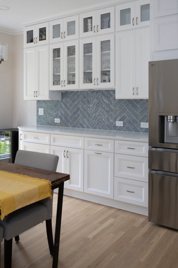 Bar cabinetry with illuminated glass uppers and herringbone tile, seen past the dining table.