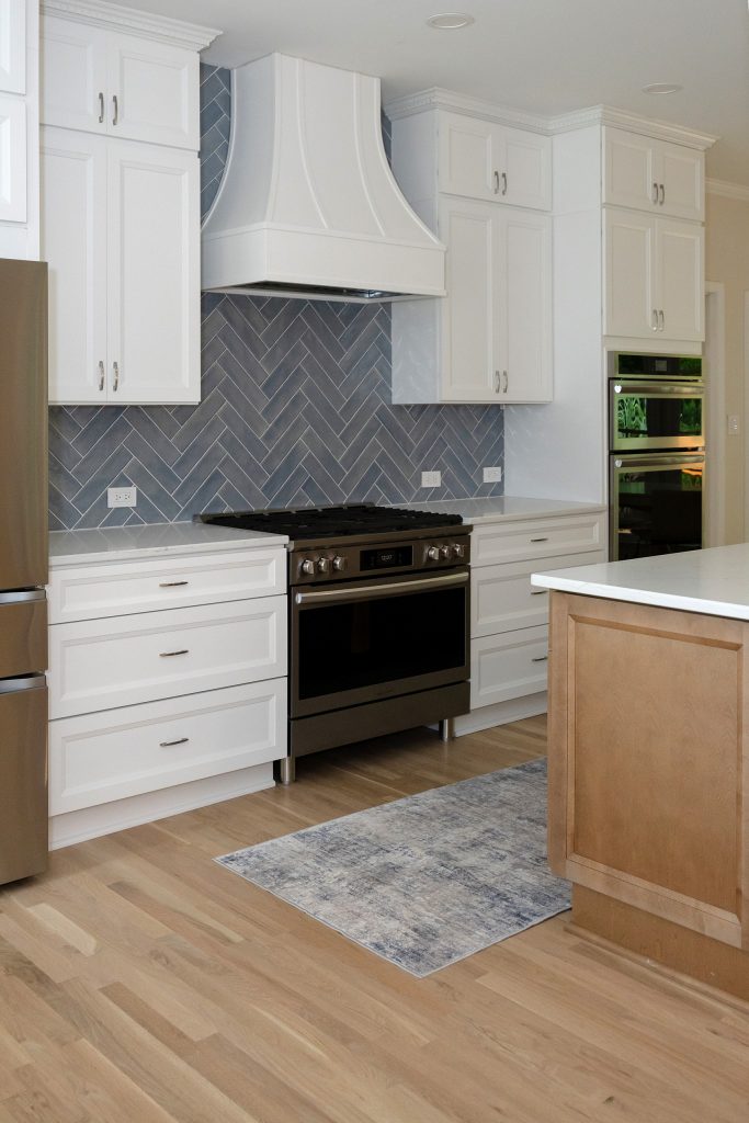 Range wall in white cabinetry with herringbone tile, beside the stained maple island and a runner rug.