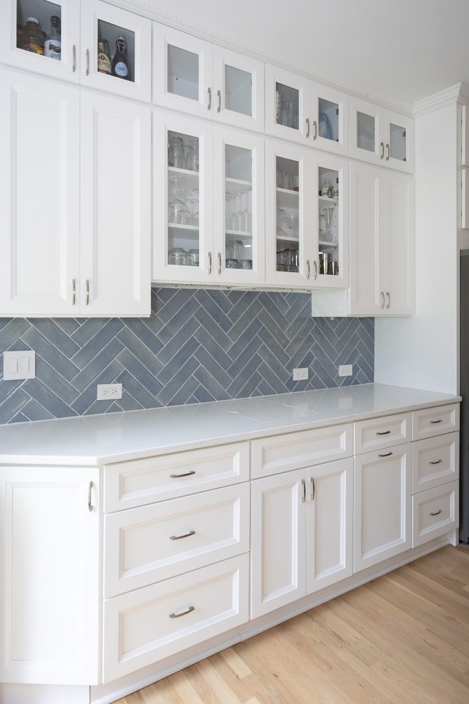 Bar cabinetry straight on, with illuminated glass-front uppers, herringbone backsplash and quartz counter.