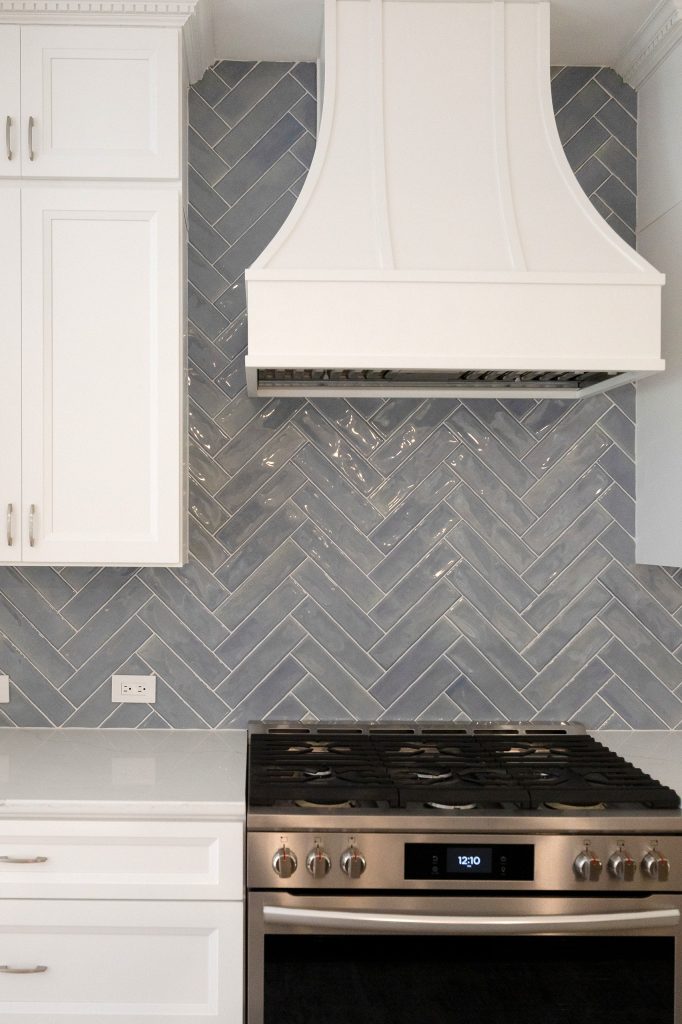 Curved white hood centred above the stainless gas range against the glossy blue-grey herringbone backsplash.