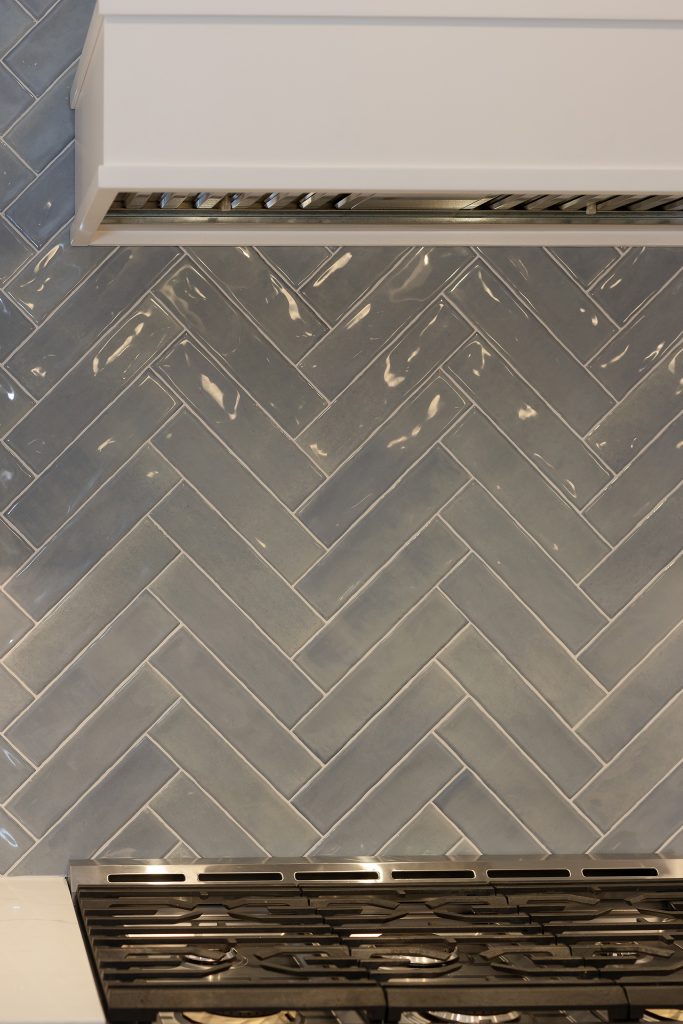 Close detail of the glossy blue-grey herringbone backsplash tile running up behind the range to the vent hood.