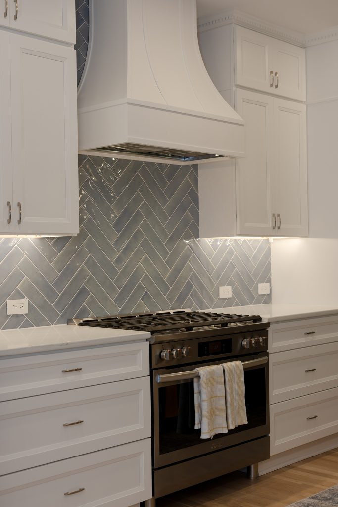Range wall with a curved white hood, herringbone tile backsplash, slate gas range and white drawer bases under quartz counters.