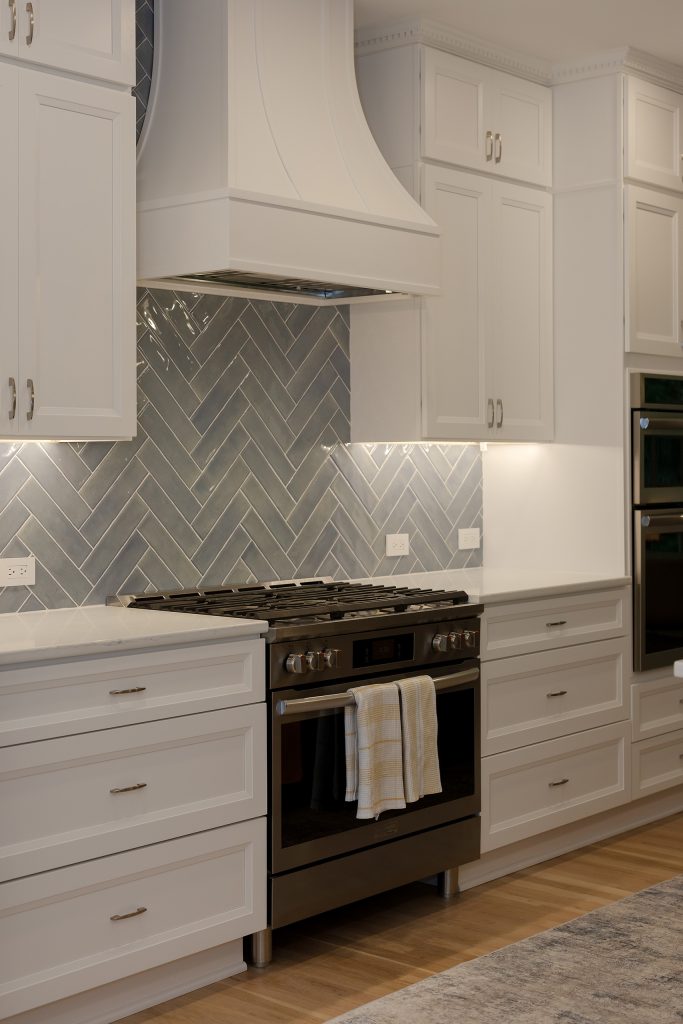 Kitchen range wall with white drawer cabinetry, herringbone backsplash and a slate gas range, with double wall ovens to the right.