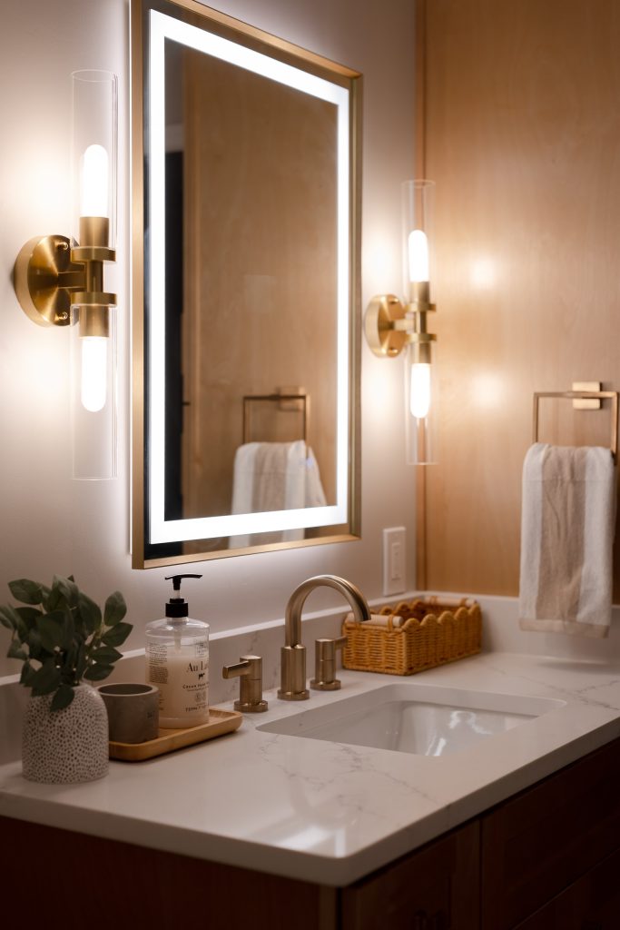 Bathroom vanity with a backlit gold-framed mirror, brushed brass sconces and faucet, white quartz counter and a maple panel wall.