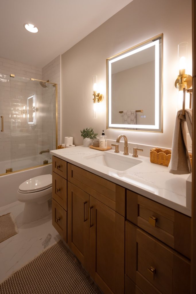 Bathroom with a glass-enclosed tub, toilet and maple vanity beneath a backlit mirror and brass sconces.