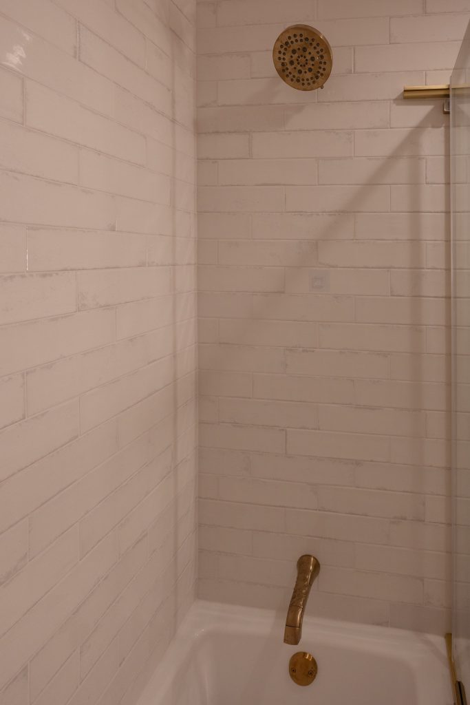Detail of the tub surround in glossy white subway tile with a brushed brass shower head and tub spout.