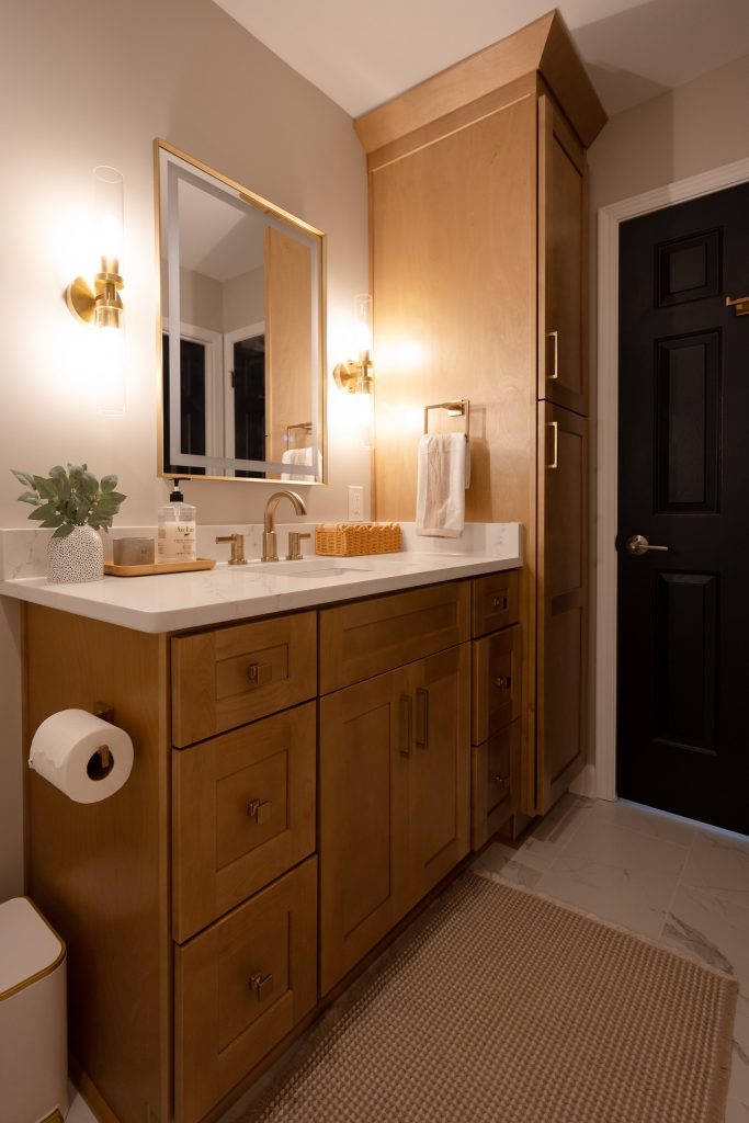 Vanity and full-height linen tower in stained maple, with marble-look porcelain flooring and a woven runner.