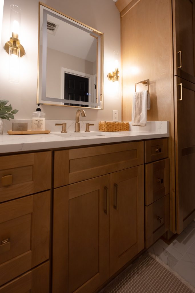 Vanity detail with a backlit mirror, brass sconces and quartz counter beside the maple linen tower.