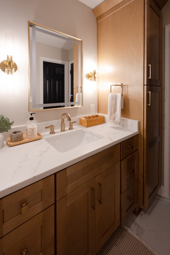 Quartz vanity top with an undermount basin and brushed brass faucet, below a backlit gold-framed mirror.
