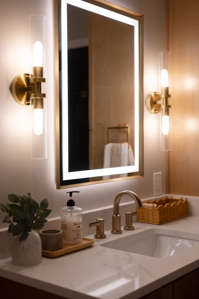 Close view of the vanity with a backlit mirror between two brass sconces above a quartz counter and undermount basin.
