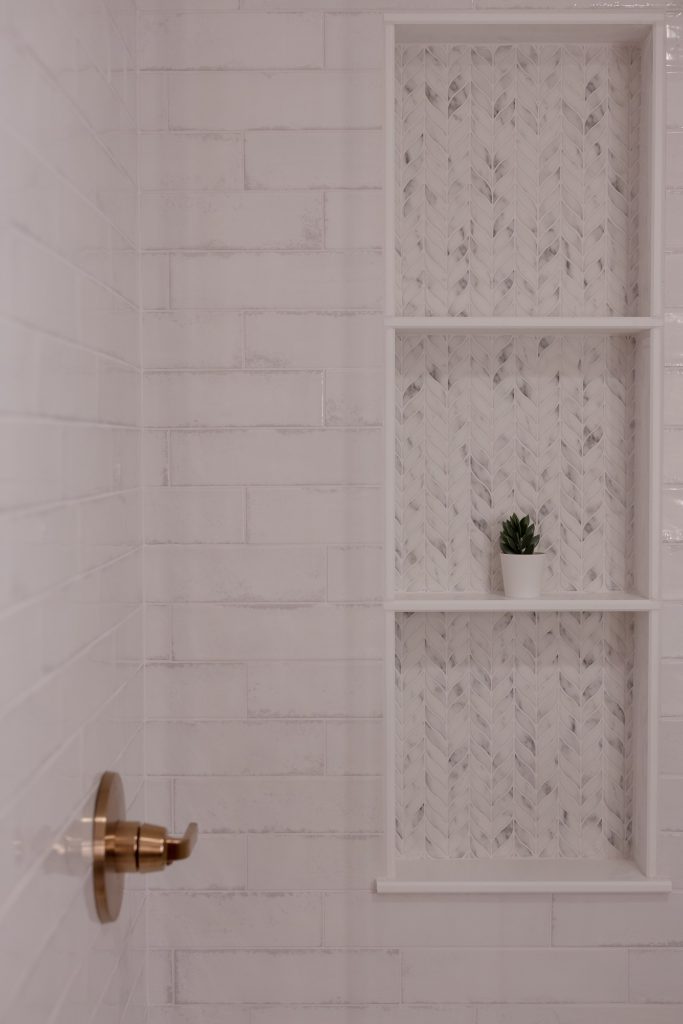 Shower wall in glossy white subway tile with a three-section niche lined in chevron-cut marble mosaic and a brushed brass valve.