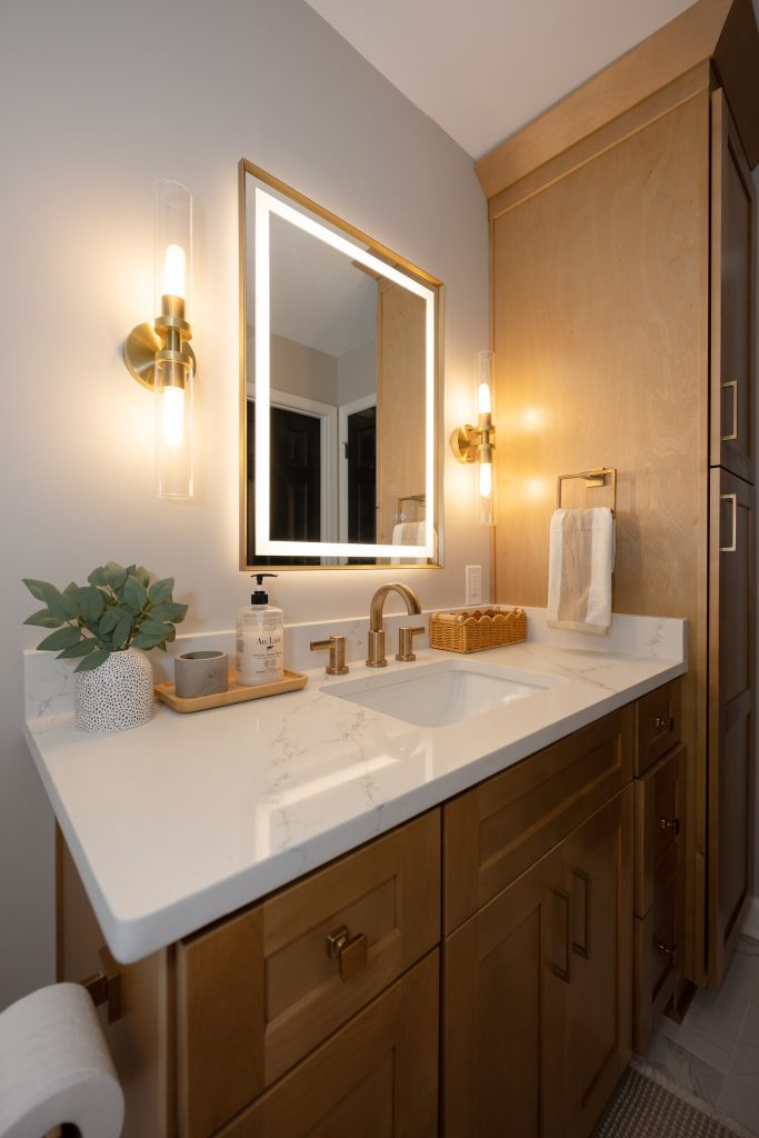 Stained maple vanity with a full-height linen tower, quartz counter, brass fittings and a backlit mirror.
