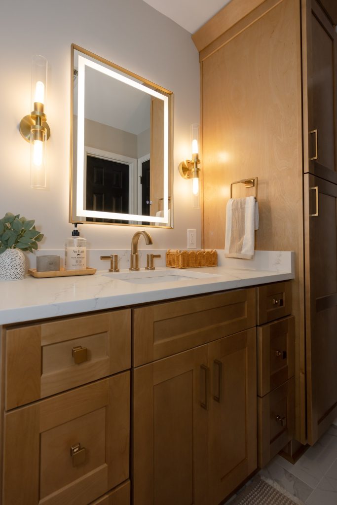 Maple vanity and linen tower under a backlit mirror, with brushed brass hardware and a white quartz top.