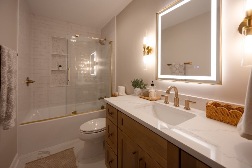 Remodeled bathroom with tub and glass enclosure, tiled niche, maple vanity and a backlit mirror between brass sconces.